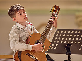 Junge spielt GItarre auf der Bühne sitzend vor ihm eiN Notenständer. Jugend musiziert, Deutscher Musikrat 2023.