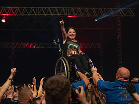 © WOA Festival GmbH JUnge Frau im Rollstuhl die von Besucherinnen eines Konzertes vor der Bühne hochgehoben wird.