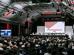 Menschen vor einer Bühne, auf der eine Versammlung abgehalten wird