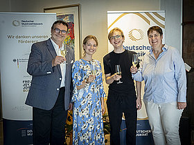 4 Personen vor Plakat Deutscher Musikwettbewerb mit Sektgläsern in der Hand. Ergebnisbekanntgabe.